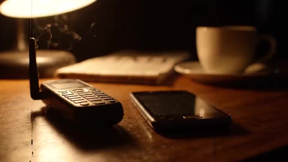 Een bureau bij nacht onder een enkele lamp. Een mobiele telefoon uit de jaren 90 met uitgeklapte antenne ligt naast een moderne smartphone, beide schermen donker. Op de achtergrond onscherp een notitieblok en een kop koffie.